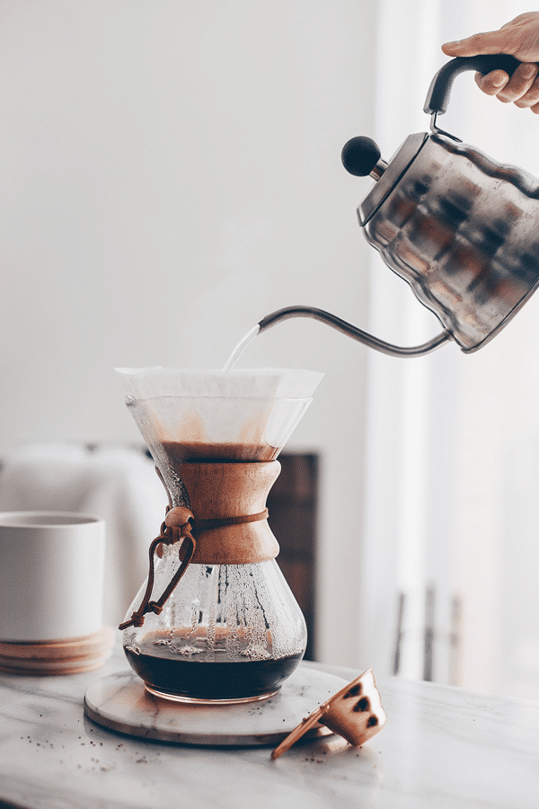 Pour-over coffee brewing with a gooseneck kettle into a Chemex on a marble countertop in a bright, modern kitchen