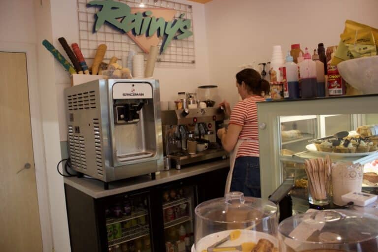 Barista preparing drinks at Rainy’s café, proudly serving Mini & Mighty Coffee alongside desserts and soft serve in a vibrant, welcoming space.