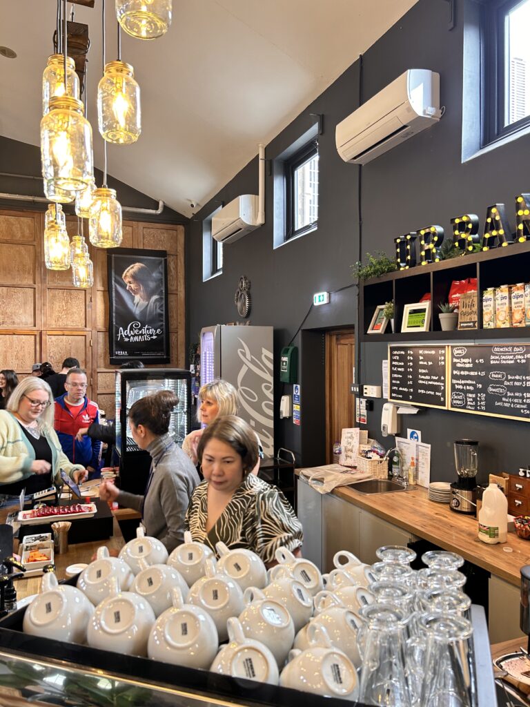 Busy café scene with customers at the counter, white mugs stacked near a barista station, and warm lighting from hanging mason jar lamps. A black feature wall displays the menu and snack shelves, with “URBAN” lit up in marquee-style lights. The atmosphere is lively and welcoming.